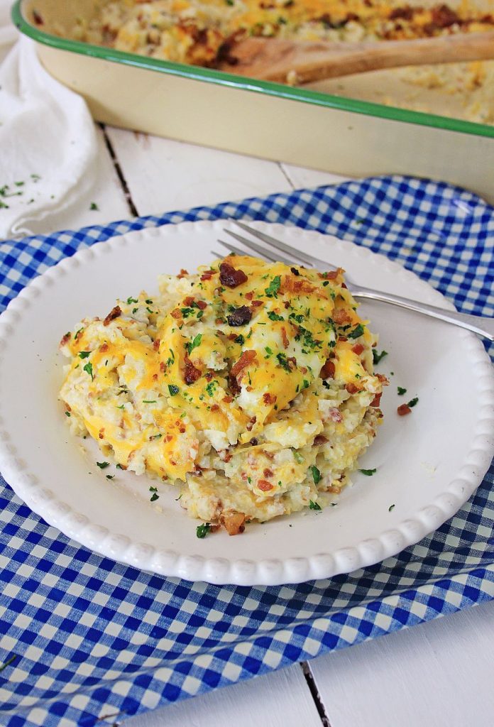 A serving of casserole on a white plate with a fork. The casserole is topped with melted cheese, crispy bacon bits, and chopped parsley. It is set on a blue checkered cloth and a casserole dish is in the background.