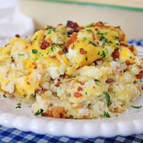 A serving of casserole topped with crumbled bacon and chopped parsley on a white plate. The dish is placed on a blue and white checkered tablecloth.