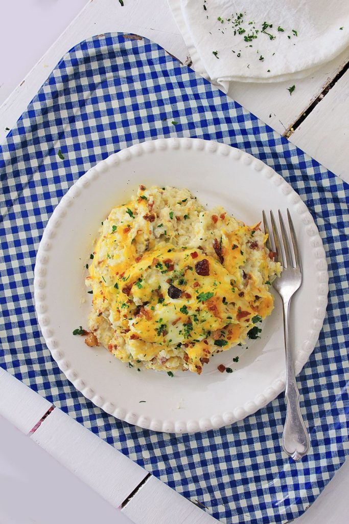 A plate of casserole topped with melted cheese and herbs on a white, textured plate. The dish is placed on a blue and white checkered cloth with a fork beside it. The casserole appears to contain cheese, bacon bits, and other ingredients.