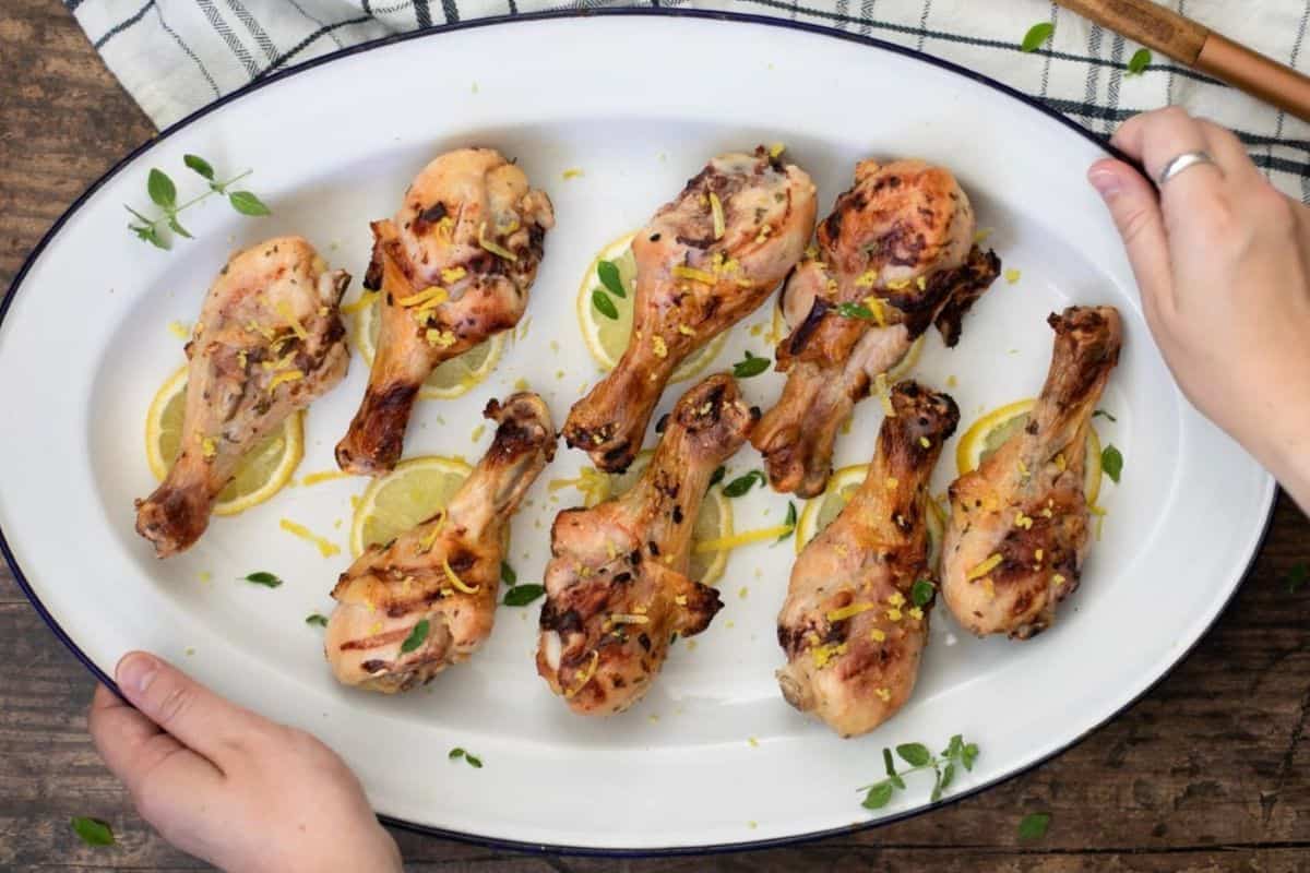 A white platter with eight cooked chicken drumsticks garnished with lemon slices, herbs, and zest. Two hands are touching the edge of the platter, and a checkered cloth is visible underneath.