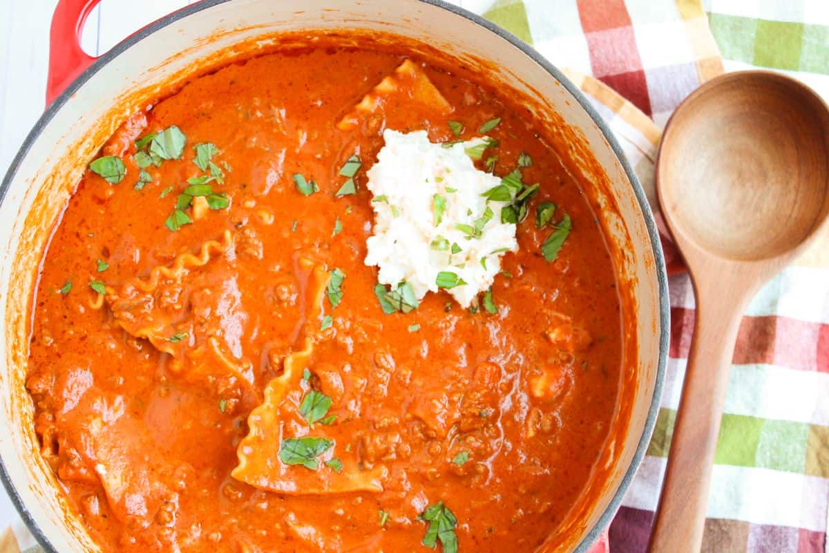 A pot of creamy lasagna soup with lasagna noodles, topped with a dollop of ricotta cheese and sprinkled with chopped herbs. A wooden spoon is beside the pot on a checkered cloth.