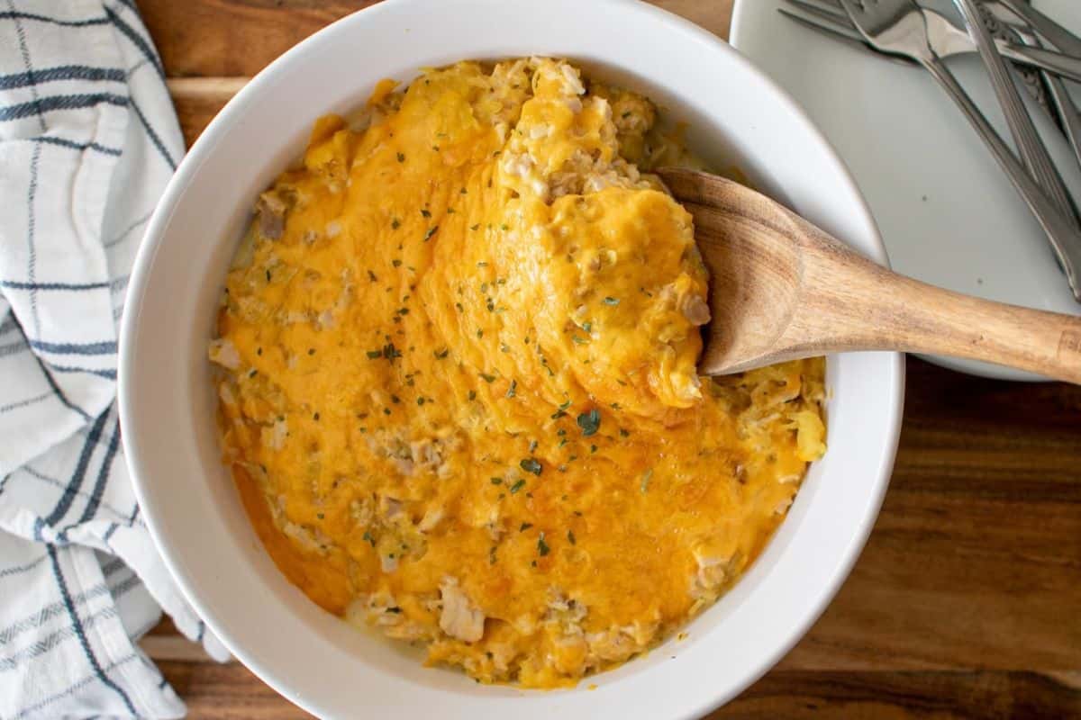 A white bowl filled with a cheesy casserole, topped with melted cheddar cheese, sits on a wooden table. A wooden spoon is scooping some out. A striped towel and stacked plates with forks are nearby.