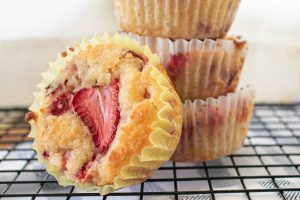 A close-up of strawberry muffins on a cooling rack. One muffin is in the foreground, featuring a visible slice of strawberry on top. A stack of three muffins is in the background, wrapped in paper liners.