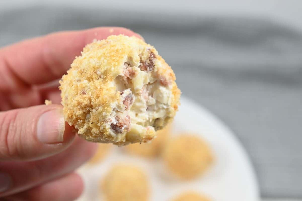 A hand holding a partially eaten ball-shaped snack with a breaded coating, revealing a filling of cream cheese, bacon, and potatoes. A white plate with similar snacks is blurred in the background.