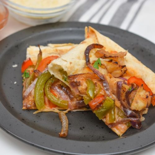 Two slices of pizza with bell peppers, onions, and chicken topping are on a black plate. In the background, there's a bowl of tomato sauce and a bowl of grated cheese on a striped cloth.