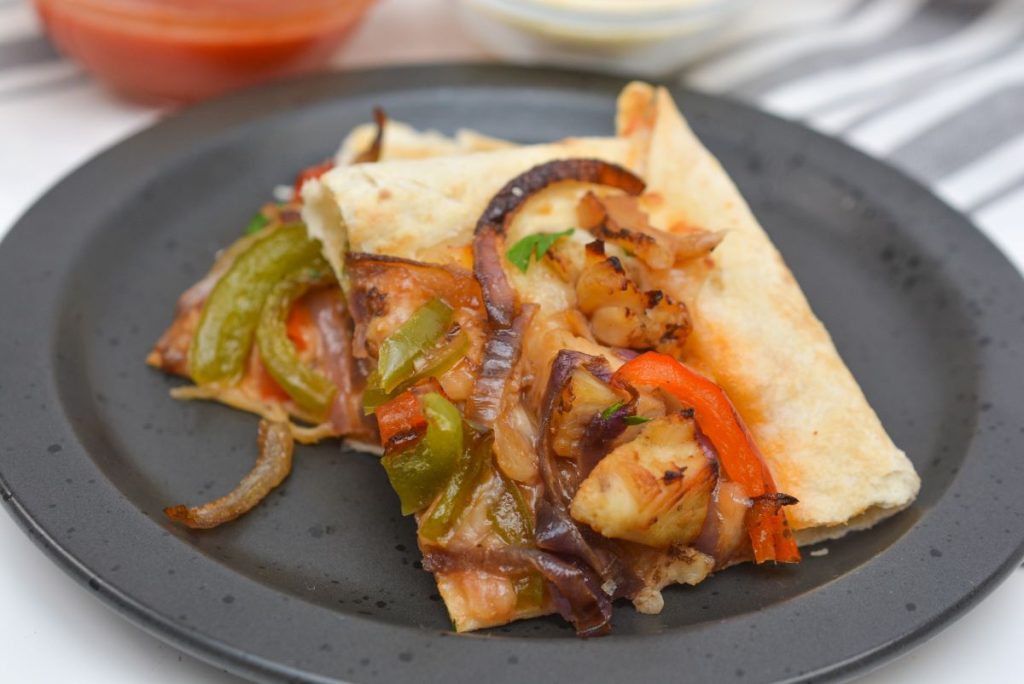 Close-up of a black plate with two slices of pizza topped with chicken, bell peppers, and onions. The crust is lightly toasted. Blurred sauce bowls are visible in the background.