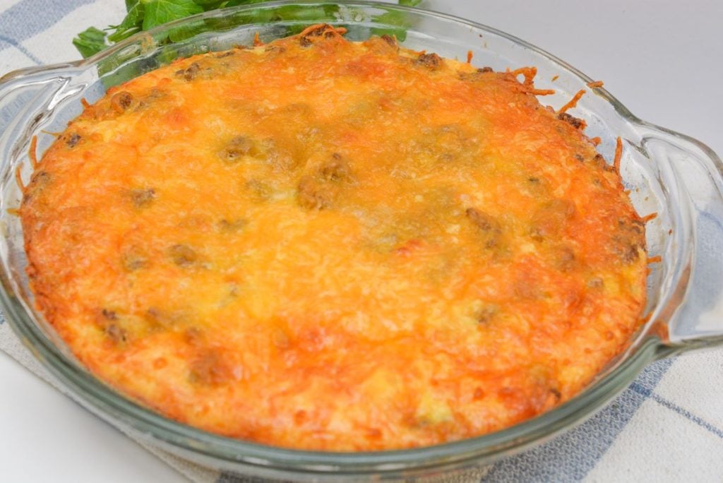 Close-up of a baked casserole in a round glass dish, featuring a golden-brown cheese topping. The dish contains visible ground meat and is placed on a folded kitchen towel. Fresh greens are partially visible in the background.