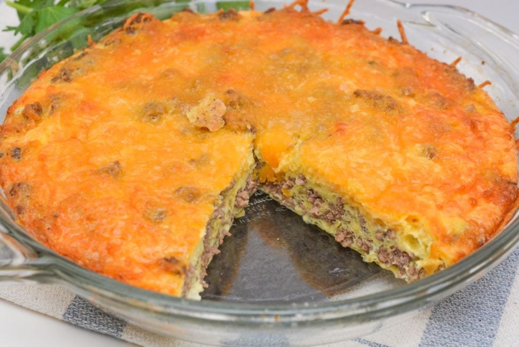 A sliced cheeseburger casserole in a glass dish, showing layers of ground beef, cheese, and egg atop a crispy, browned cheese topping. It rests on a woven cloth with greenery partially visible in the background.