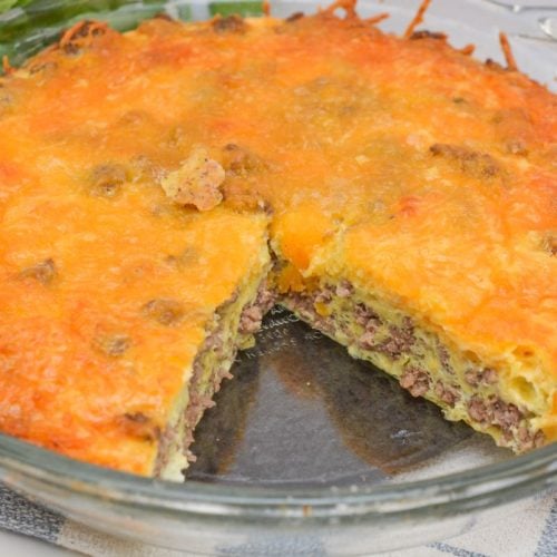 A sliced cheeseburger casserole in a glass dish, showing layers of ground beef, cheese, and egg atop a crispy, browned cheese topping. It rests on a woven cloth with greenery partially visible in the background.