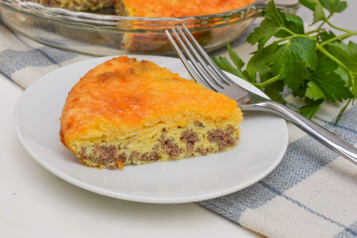 A slice of savory pie with a golden, cheesy crust and a filling likely containing ground meat sits on a white plate. A fork is placed next to it. Fresh parsley lies nearby, and the rest of the pie is visible in the glass dish behind.