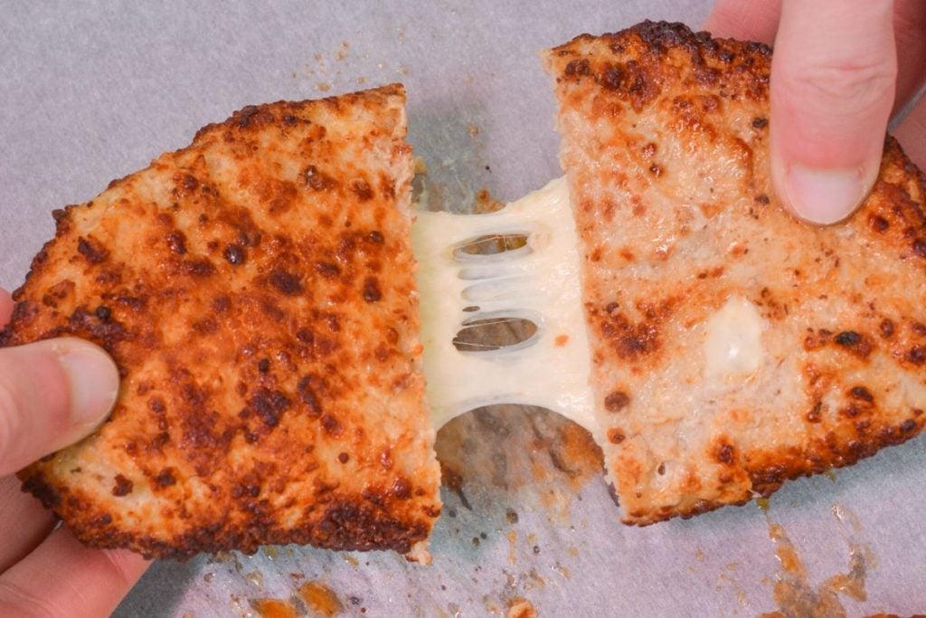 Two hands pulling apart a crispy, golden-brown breaded snack revealing melted, stretchy cheese inside, set on a parchment paper surface.