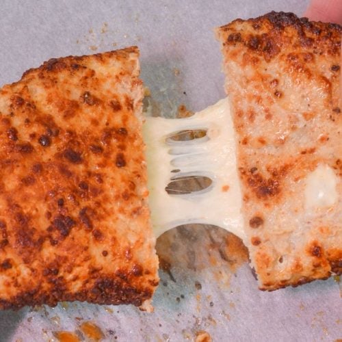 Two hands pulling apart a crispy, golden-brown breaded snack revealing melted, stretchy cheese inside, set on a parchment paper surface.