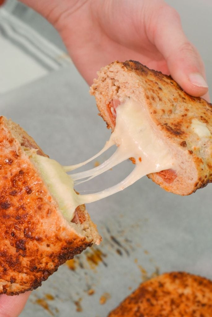 A person pulls apart a toasted sandwich revealing melted cheese stretching between the slices. The bread appears crispy and golden brown. The background is a blurred kitchen setting.