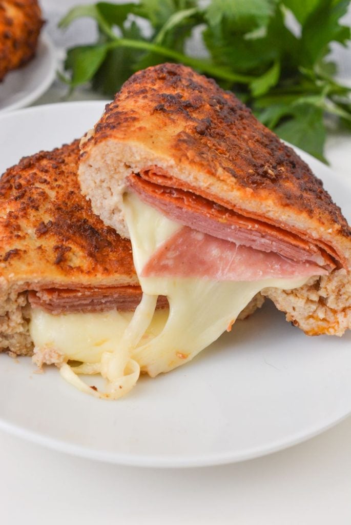 A close-up of a stuffed bread roll sliced open, revealing layers of melted cheese, ham, and salami. The crust is golden and slightly crispy, and fresh green herbs are blurred in the background.
