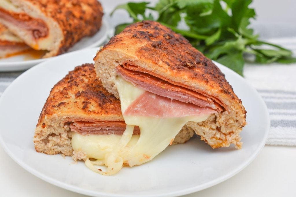 A close-up of a toasted sandwich on a white plate. The sandwich is cut in half, revealing melted cheese and slices of cured meat inside. Fresh parsley is blurred in the background.
