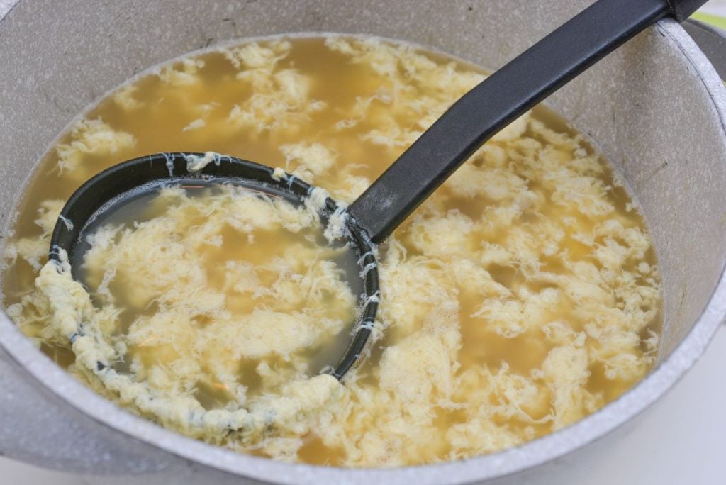 A pot of egg drop soup with a ladle. The soup features a light, translucent broth with cooked egg strands floating on the surface. The ladle is positioned partially submerged in the soup.