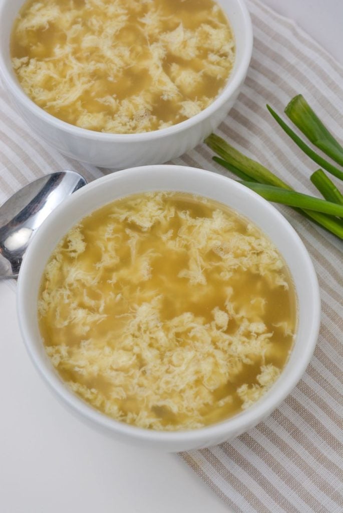 Two bowls of egg drop soup with a light broth and threads of egg sit on a striped cloth. A shiny spoon and green onions are nearby, adding a touch of color to the scene.