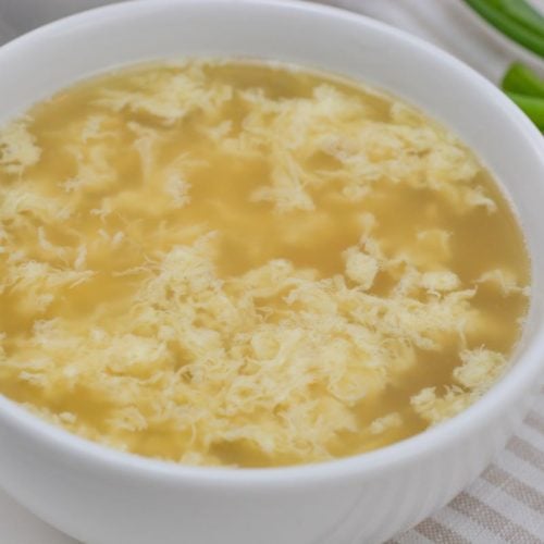 A white bowl filled with egg drop soup on a striped tablecloth. The soup has a light broth with visible wisps of egg. Green onions are blurred in the background.