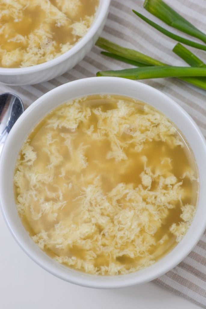 A close-up of a bowl of egg drop soup on a striped cloth. The soup features wispy egg strands in a light broth. A spoon and green onion stalks are visible nearby.