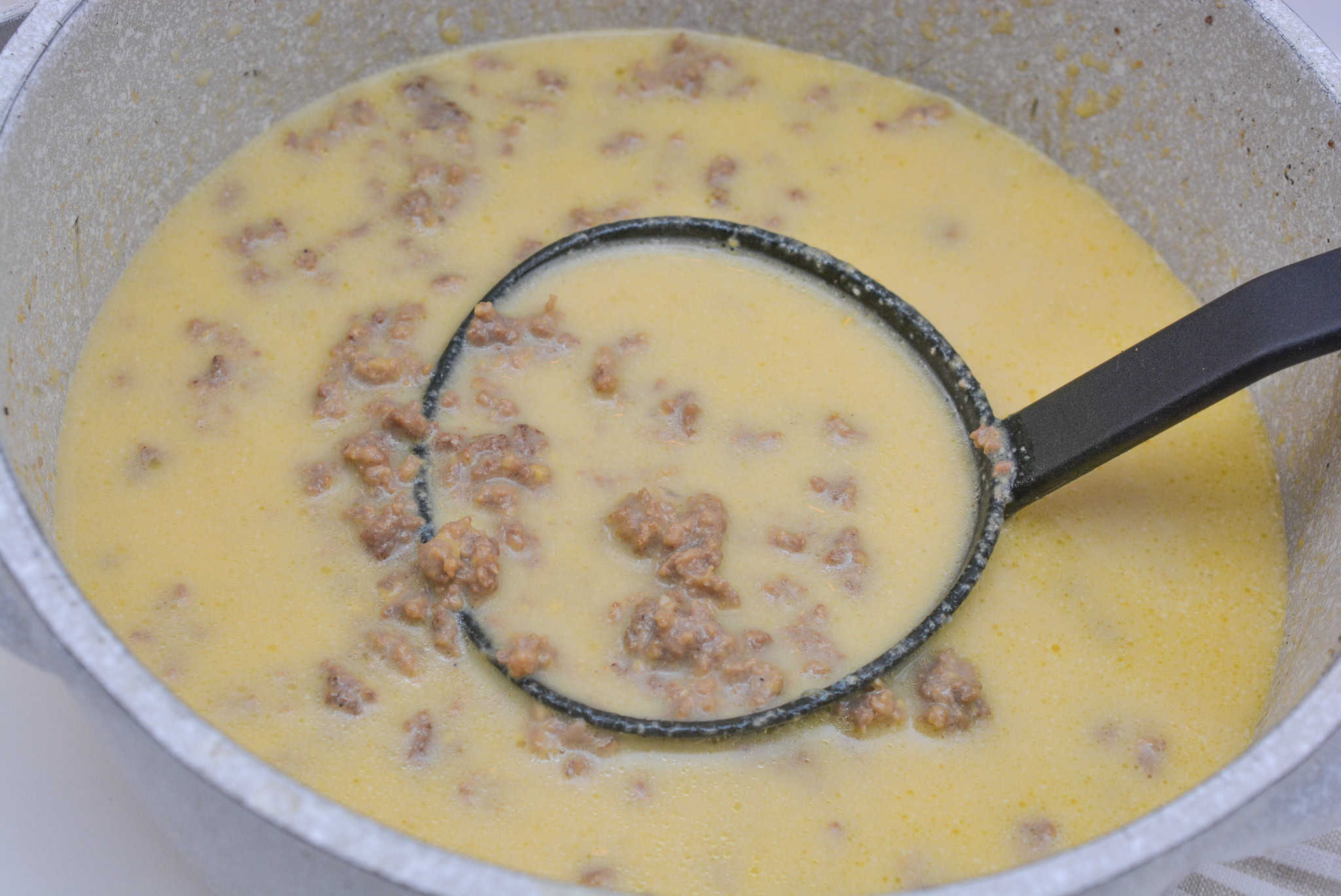 A pot of yellow cheese soup with cooked ground beef visible. A black ladle is scooping some of the soup, showing pieces of beef mixed throughout. The pot is on a white surface.