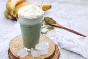 A tall glass of green smoothie topped with whipped cream and a straw, set on a wooden coaster with ice cubes. In the background, bananas and a wooden spoon with green powder are visible on a marble surface.