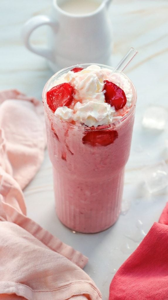 A tall glass of strawberry milkshake topped with whipped cream and strawberry slices. A clear straw is in the drink. A pitcher is in the background on a marble surface. Pink and red cloth napkins are placed nearby.