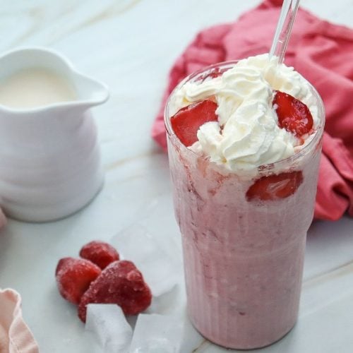 A glass of strawberry milkshake topped with whipped cream and sliced strawberries, accompanied by a small jug of milk. Frozen strawberries and ice cubes are placed nearby on a light surface with a pink cloth in the background.