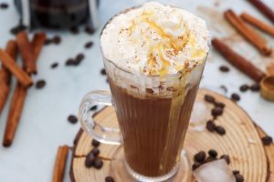Tall glass of iced coffee topped with whipped cream and caramel drizzle, set on a wooden coaster. Surrounded by scattered coffee beans, ice cubes, and cinnamon sticks, creating a cozy, inviting atmosphere.