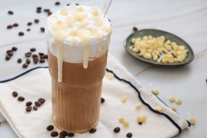 A tall glass of iced coffee topped with whipped cream and white chocolate drizzle. Coffee beans and white chocolate chips are scattered around. A plate with more white chocolate chips is in the background.