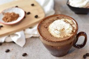 A ceramic mug filled with hot chocolate topped with whipped cream sits on a table. Coffee beans are scattered around, and a small plate with cocoa powder is on a wooden board in the background.