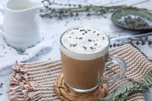 A glass mug filled with coffee topped with frothy milk and lavender sprinkles sits on a braided coaster. Next to it is a fringed cloth and a plate with more lavender. A white pitcher is blurred in the background.