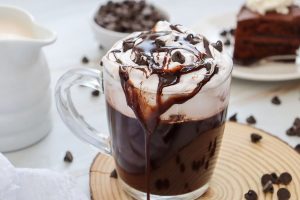 A glass mug filled with hot chocolate, topped with whipped cream, chocolate syrup, and chocolate chips. It rests on a wooden coaster, with chocolate chips scattered around. A slice of chocolate cake is blurred in the background.