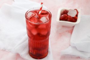 A tall glass filled with red iced drink and ice cubes, featuring a red and white striped straw. The background has a white cloth, and there is a small dish with fresh strawberries and ice cubes. The setting is on a light pink surface.