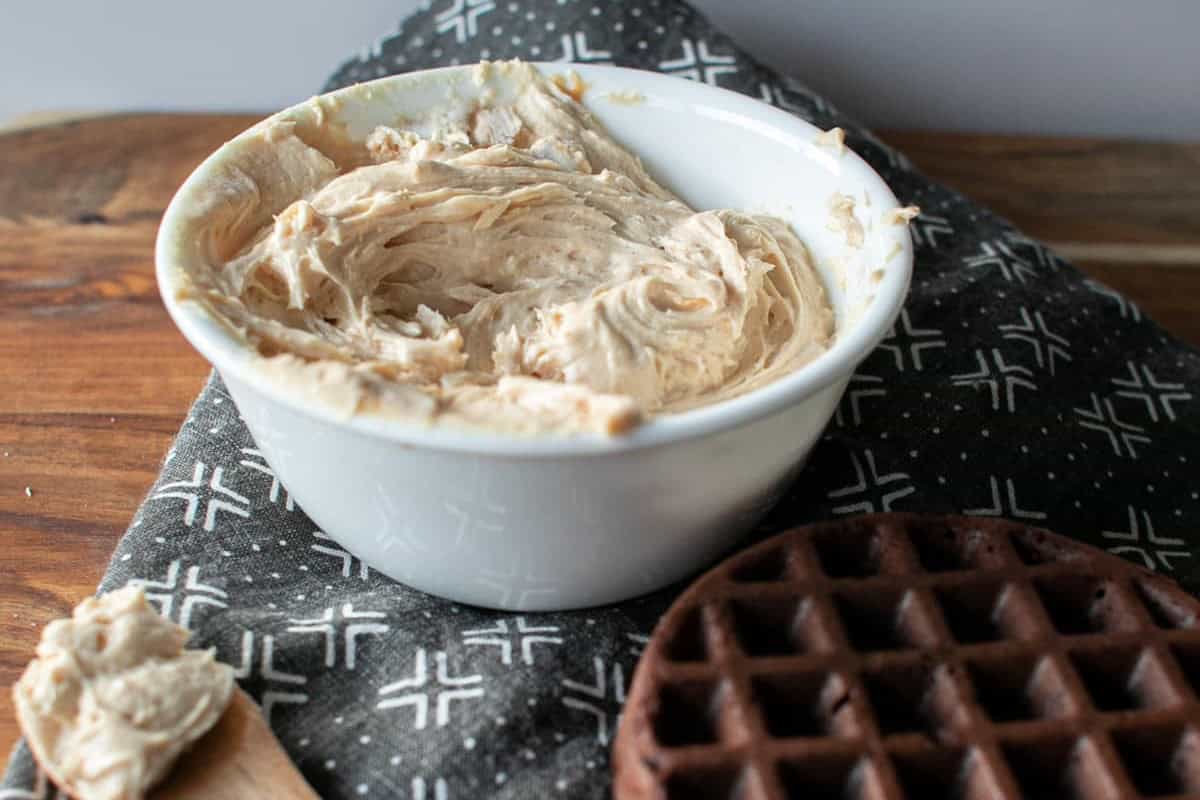 A white bowl filled with creamy spread sits on a dark-patterned cloth. A wooden spoon with a bit of spread rests nearby, and a chocolate waffle lies beside them on a wooden surface.