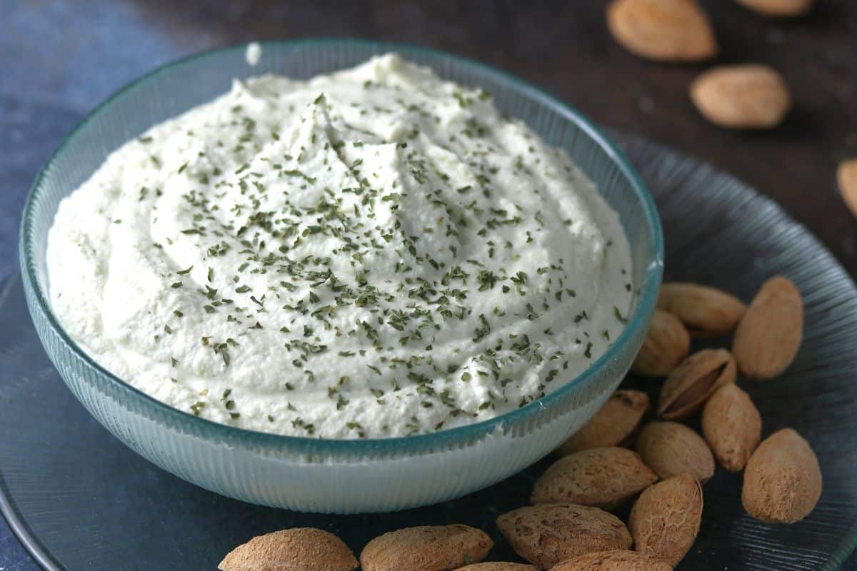 A bowl of creamy dip topped with chopped herbs, surrounded by almonds, sits on a dark surface.