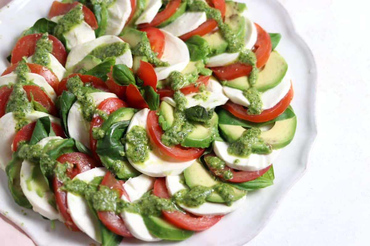 A fresh Caprese salad on a white plate showcases alternating slices of tomatoes, mozzarella, and avocado, garnished with basil leaves. The salad is drizzled with a green pesto dressing.