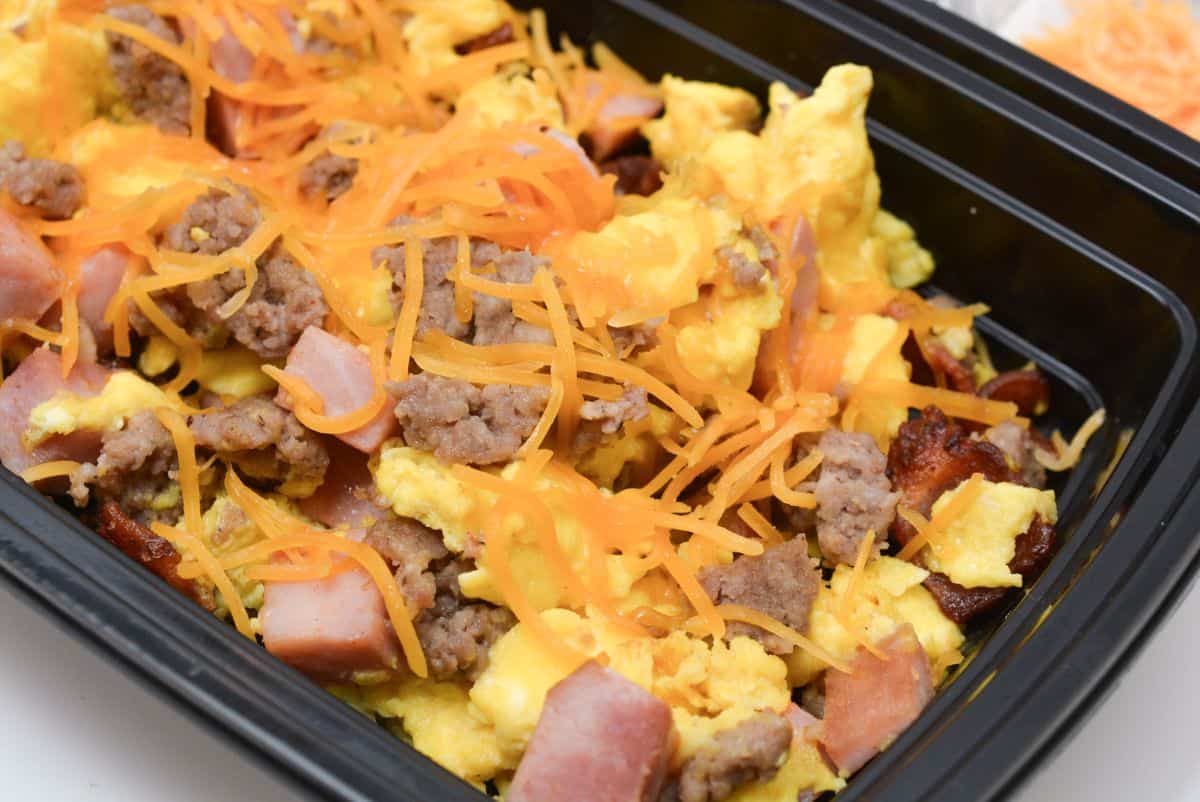 A black container filled with scrambled eggs, chunks of ham, sausage crumbles, and shredded cheddar cheese.