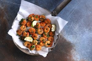 A pan filled with cooked shrimp garnished with chopped herbs and lemon wedges, resting on a piece of parchment paper. The pan is placed on a textured, rustic surface.
