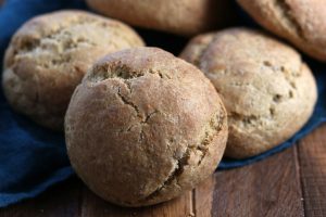 Close-up of several round, golden-brown bread rolls with cracked tops, resting on a dark blue cloth on a wooden surface. The rolls appear freshly baked and rustic in appearance.