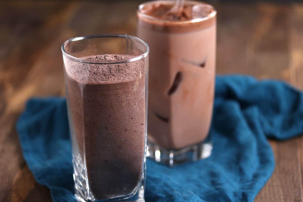Two tall glasses filled with chocolate milkshake stand on a wooden surface. One shake is topped with chocolate powder, while the other, slightly blurred, has ice cubes. A dark blue cloth is casually draped beneath the glasses.