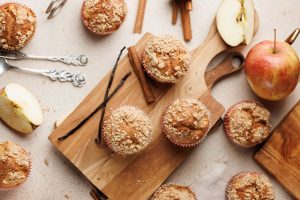 Freshly baked muffins with crumb topping on wooden boards, surrounded by sliced apples, whole apples, cinnamon sticks, and antique silverware on a beige surface.