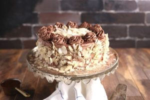 A chocolate cake decorated with chocolate swirls on top and coated with sliced almonds on the sides sits on a vintage cake stand. The background features a rustic brick wall, and a folded white cloth is underneath the stand.