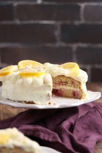 A delectable cake topped with creamy white frosting and lemon slices is displayed on a white cake stand. The cake has visible layers with a raspberry filling. A purple cloth is placed underneath, and a dark brick wall forms the background.