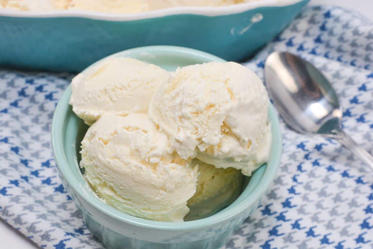 A light blue bowl filled with three scoops of vanilla ice cream sits on a patterned cloth next to a metal spoon and a larger dish of ice cream in the background.