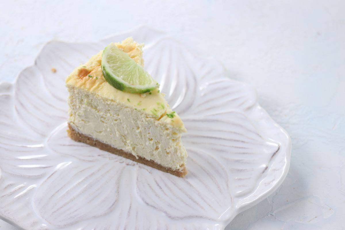 A slice of key lime cheesecake with a graham cracker crust, topped with a lime wedge, served on a white, flower-shaped plate against a light background.