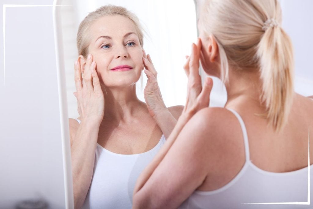 A middle-aged woman in a white tank top stands in front of a mirror, gently touching her face and smiling softly as she examines her reflection—curious about using peptides for beginners in her skincare routine.