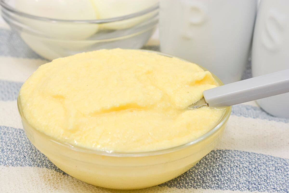 A clear glass bowl filled with creamy, yellow grits sits on a striped cloth. A spoon rests in the bowl. Out-of-focus containers and another glass bowl appear in the background.