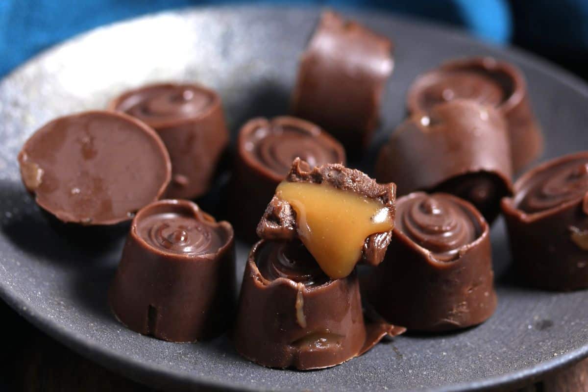 A plate of round chocolate candies, some cut open to reveal gooey caramel filling inside, displayed on a dark dish.
