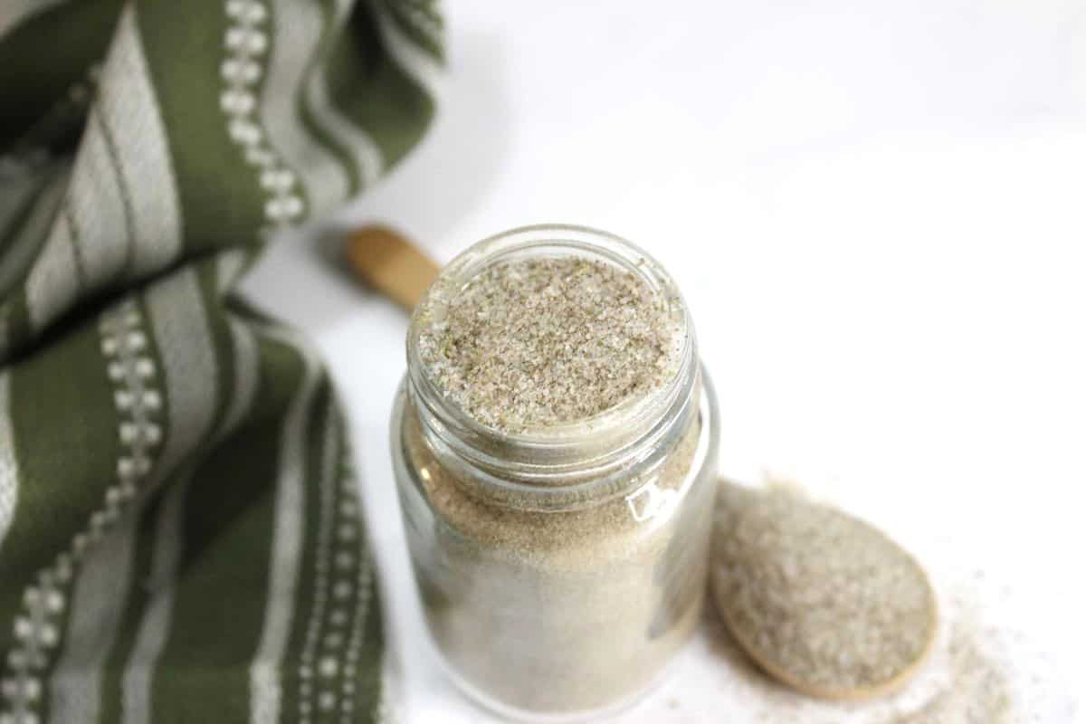 A glass jar filled with seasoned salt sits on a white surface, next to a wooden spoon holding some of the salt. A green and white striped cloth is partially visible in the background.