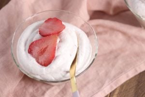 A glass bowl filled with creamy whipped dessert topped with two strawberry slices, placed on a pink cloth with a gold spoon resting inside the bowl.